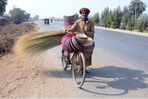A roadside vendor walks along Muzaffargarh Road with his bicycle loaded with handmade household items, including bread baskets and brooms, offering them for sale to passersby.