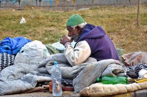 In the cold weather, an elderly homeless man sits by the roadside, wrapped in blankets and sipping a warm drink, quietly highlighting the hardships faced by marginalized communities and the gaps in social support systems.
