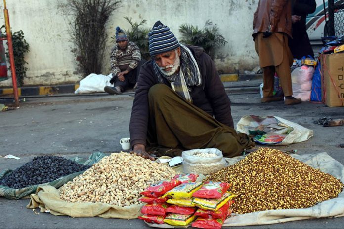 An elderly vendor sells peanuts and dry fruits on the roadside to attract passersby and earn a living in the local market