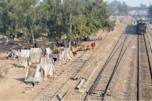 Nomadic families, including women and children, stand near their makeshift shanties built along the railway tracks, highlighting the hardships and challenges faced by marginalized communities.