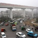 Vehicles pass along the road as the Orange Line train moves in the background amid morning fog at Thokar Niaz Baig in the Provincial Capital.