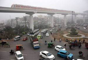Vehicles pass along the road as the Orange Line train moves in the background amid morning fog at Thokar Niaz Baig in the Provincial Capital.