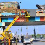 HMC workers are busy installing an LED screen for advertisements on the bypass bridge, using heavy machinery on Jamshoro Road
