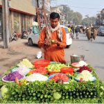 A roadside vendor prepares fresh vegetable salad at his cart along a busy city street, attracting customers with neatly arranged seasonal vegetables on a sunny afternoon