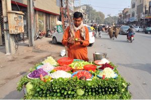 A roadside vendor prepares fresh vegetable salad at his cart along a busy city street, attracting customers with neatly arranged seasonal vegetables on a sunny afternoon