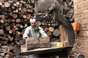 An elderly woodcutter chops a log into pieces for firewood while standing at a timber warehouse in the city.