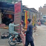 A person sitting on the rear seat of a motorcycle carries a purchased charpai from a shop