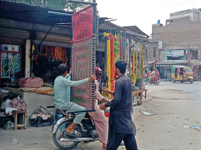 A person sitting on the rear seat of a motorcycle carries a purchased charpai from a shop