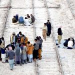 People are spending their free time playing cards on the railway tracks near the railway station, with the people standing around them watching.