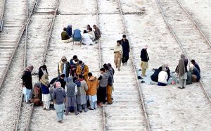 People are spending their free time playing cards on the railway tracks near the railway station, with the people standing around them watching.