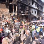 Security personnel stand as rescue workers search the debris for fire victims at Gul Plaza.