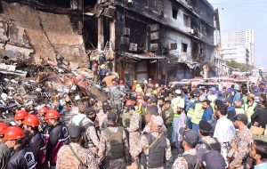 Security personnel stand as rescue workers search the debris for fire victims at Gul Plaza.