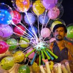 A street vendor sells illuminated balloons at a busy location.