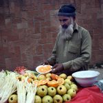 An elderly vendor sells fresh vegetables and fruit salad on his bicycle displayed for sale at a roadside