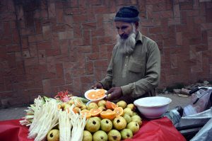 An elderly vendor sells fresh vegetables and fruit salad on his bicycle displayed for sale at a roadside