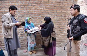 A police officer standing in an alert position while a health worker administers a polio vaccine to a child during a door-to-door vaccination campaign in Dalazak Road Faqirabad area.