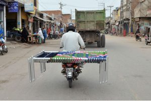 A motorcyclist carries a traditional charpai (woven bed) on his bike while passing through a busy city road.