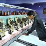 A worker supervises embroidery design work on fabric using a computerized embroidery machine at a local factory.