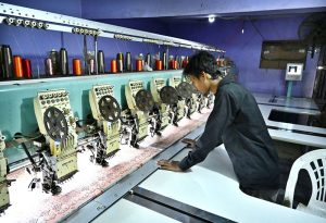 A worker supervises embroidery design work on fabric using a computerized embroidery machine at a local factory.