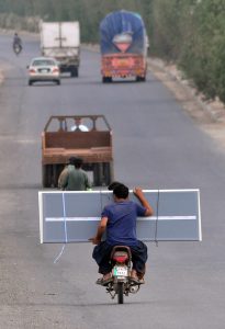 A man seated on the rear of a motorcycle holds a PVC door panel as they move along a city road.