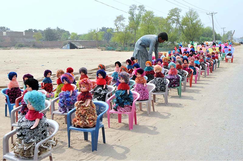 A vendor arranges handmade colorful dolls on chairs along a roadside to attract customers