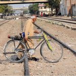 An elderly man pushes his bicycle while crossing the railway tracks as a train approaches in the background, highlighting safety risks and daily commuting challenges in the city
