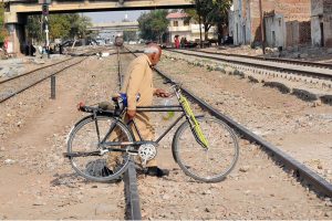 An elderly man pushes his bicycle while crossing the railway tracks as a train approaches in the background, highlighting safety risks and daily commuting challenges in the city