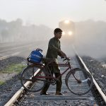 A young boy pushes a bicycle carrying a gas cylinder while crossing a railway track, while a train approaching in the background, highlighting safety risks and daily commuting challenges