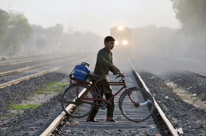 A young boy pushes a bicycle carrying a gas cylinder while crossing a railway track, while a train approaching in the background, highlighting safety risks and daily commuting challenges
