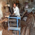 A woodworker guides a slab of raw timber through a surface planer in a local carpentry shop, where raw materials are transformed into finished products for the domestic market