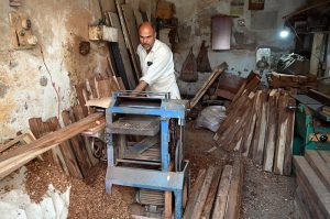 A woodworker guides a slab of raw timber through a surface planer in a local carpentry shop, where raw materials are transformed into finished products for the domestic market