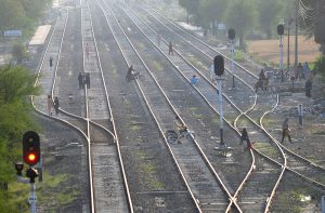People cross and walk along multiple railway tracks while motorcyclists carefully navigate through the lines at a railway yard, despite safety risks.