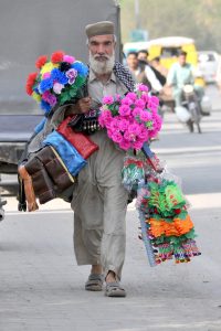 A street hawker carrying colorful artificial flowers and decorative items walks along Vehari Road in search of customers