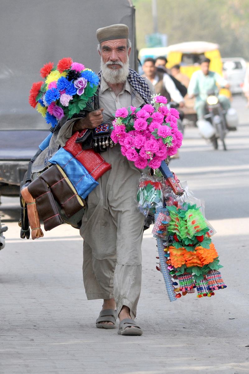 A street hawker carrying colorful artificial flowers and decorative items walks along Vehari Road in search of customers