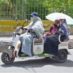 A family travels on a three-wheeled scooter, using an umbrella for shade to protect themselves from the scorching heat in the city