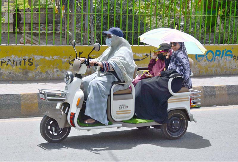 A family travels on a three-wheeled scooter, using an umbrella for shade to protect themselves from the scorching heat in the city