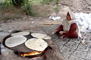 A woman prepares traditional roti on a clay stove using firewood at an outdoor rural setting.