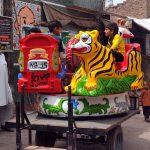 A local street vendor operates a hand-painted, cart-mounted kiddie ride, offering affordable entertainment for children in a bustling neighborhood. These mobile carousels, decorated with vibrant folk art, remain a cherished part of street life