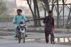 Two youngsters struggle onward after their motorcycle runs out of fuel, with one riding while the other tows the bike along a city road using a scarf.