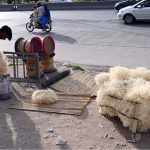 A roadside vendor arranges and prepares husk for room coolers, along with household items and scrubbers for sale beside a busy city road as traffic passes by