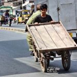 A youngster holding cart while sitting on rear seat of motorcycle at Millat Road