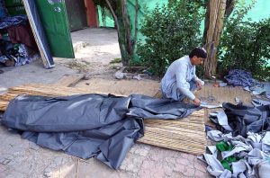 A roadside vendor arranges and prepares husk for room coolers, along with household items and scrubbers for sale beside a busy city road as traffic passes by