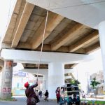 A worker paints the underside of a flyover using a long roller while traffic moves below on the roadside during ongoing developing project in the city