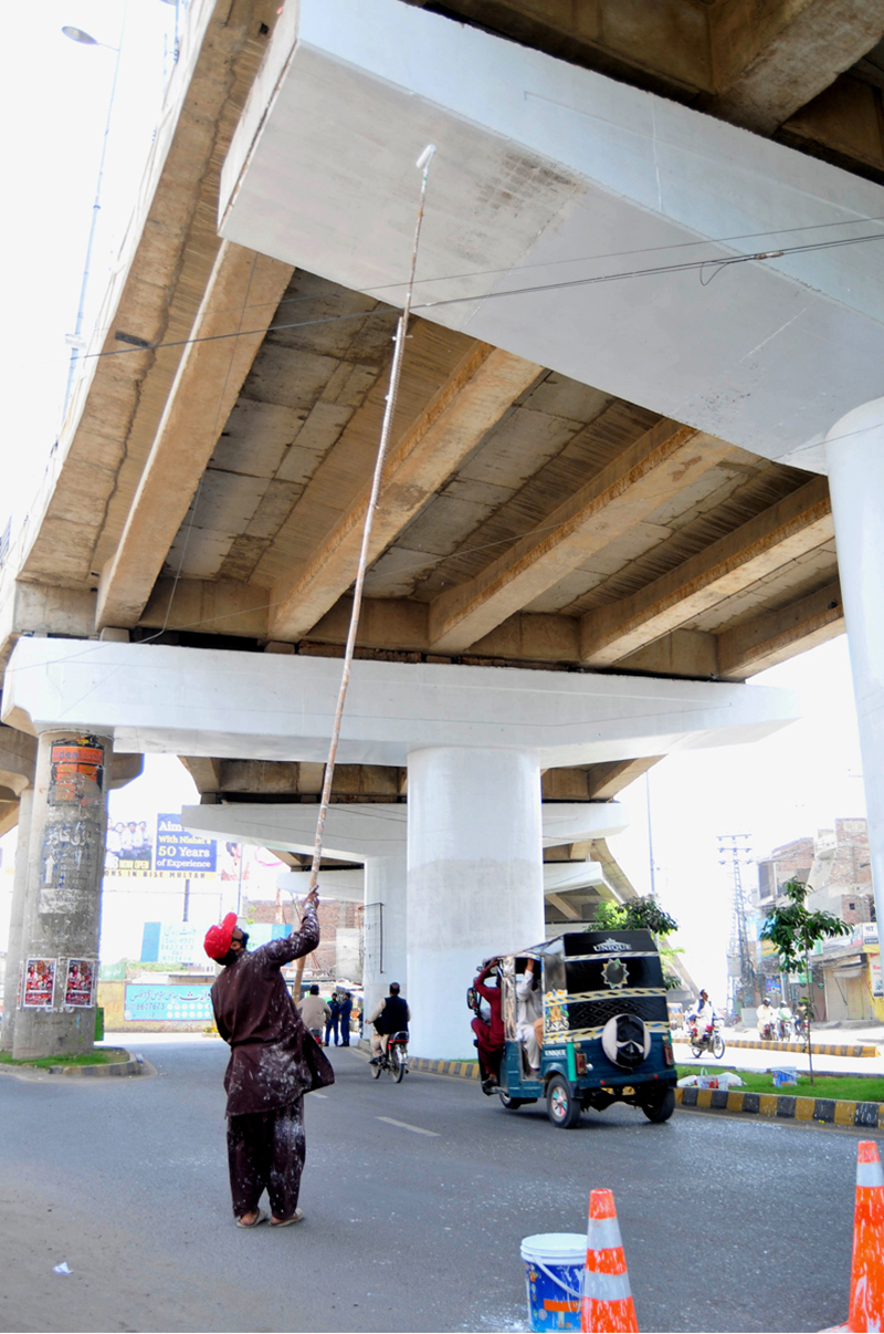 A worker paints the underside of a flyover using a long roller while traffic moves below on the roadside during ongoing developing project in the city