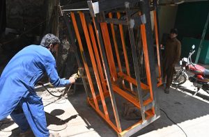 A worker sprays paint on a big size metal frame at a workshop while another looks on in the background.