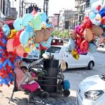A roadside woman vendor sits with a child beside a cart, surrounded by colorful helium balloons, including character-themed and animal-shaped designs, waiting for customers along a busy city street IN THE Commercial Market area