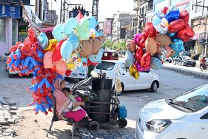 A roadside woman vendor sits with a child beside a cart, surrounded by colorful helium balloons, including character-themed and animal-shaped designs, waiting for customers along a busy city street IN THE Commercial Market area