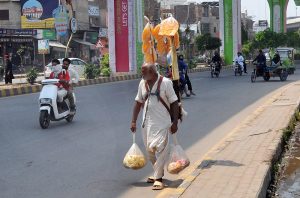 An elderly street vendor walks along a busy road carrying snacks and goods on his shoulders, heading toward customers amid the city’s daily hustle.