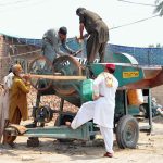 Workers repair and maintain a traditional agricultural threshing machine at a roadside workshop, preparing equipment for the harvesting season