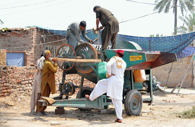 Workers repair and maintain a traditional agricultural threshing machine at a roadside workshop, preparing equipment for the harvesting season